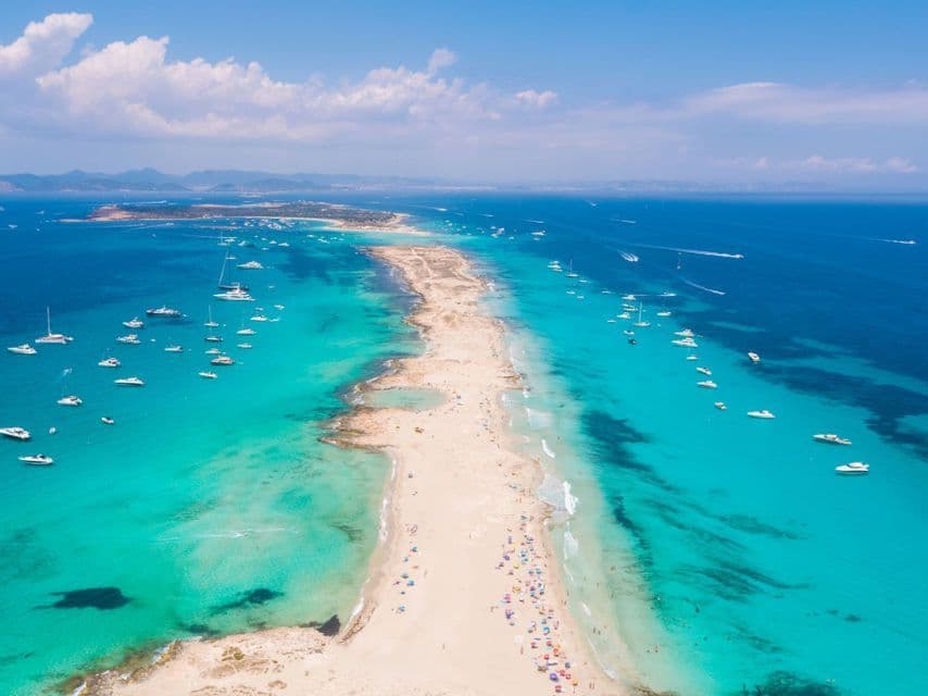Una vista aerea di una lunga lingua di sabbia che si estende in un mare turchese, con molte barche ancorate e persone sulla spiaggia.