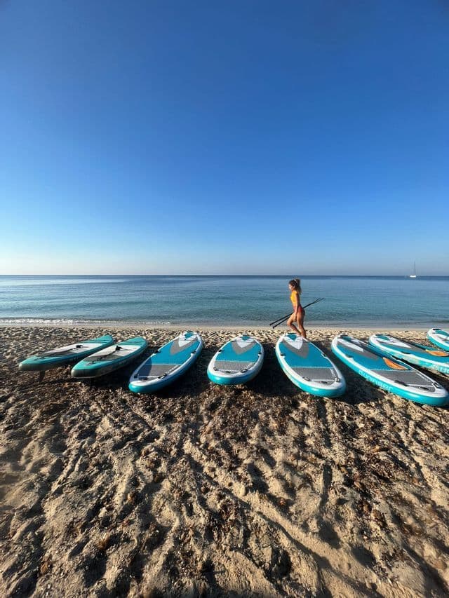 Una donna con una pagaia cammina su una spiaggia sabbiosa, superando diverse tavole da paddleboard allineate in riva al mare.
