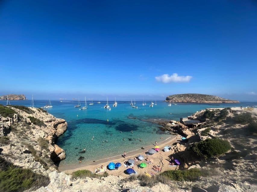Una vista dall'alto di una caletta sabbiosa con acqua turchese, ombrelloni colorati sulla spiaggia e barche a vela ancorate nella baia.