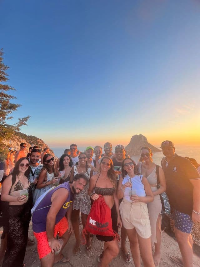 Un gruppo WeRoad in viaggio posa per una foto su una scogliera a picco sul mare e su un isolotto roccioso al tramonto.