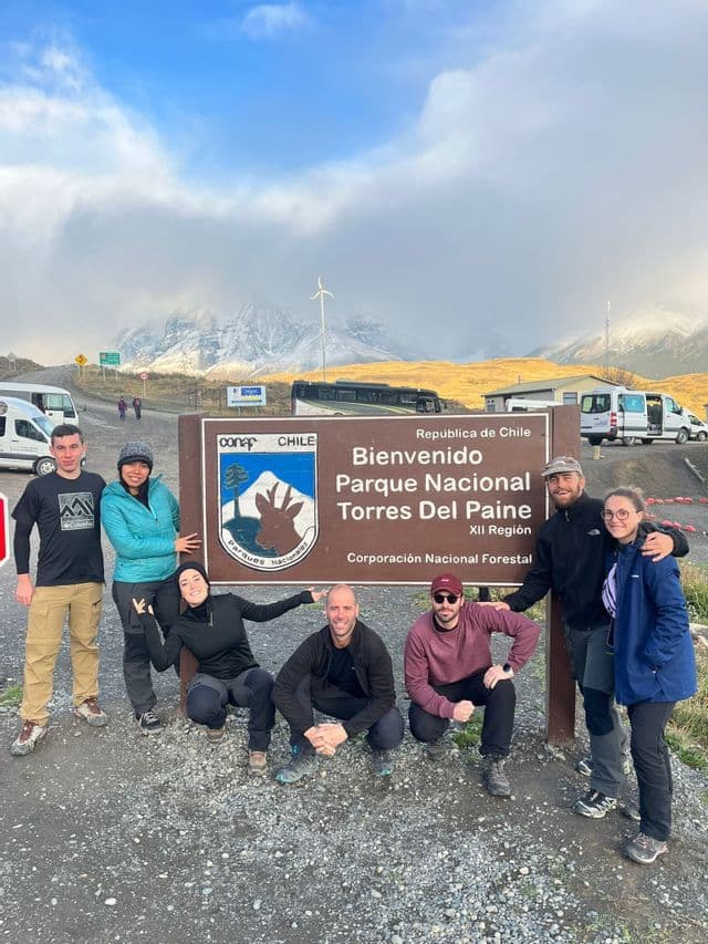 A WeRoad group trip poses together for a photo at a national park entrance sign, with misty, snow-capped mountains behind them.