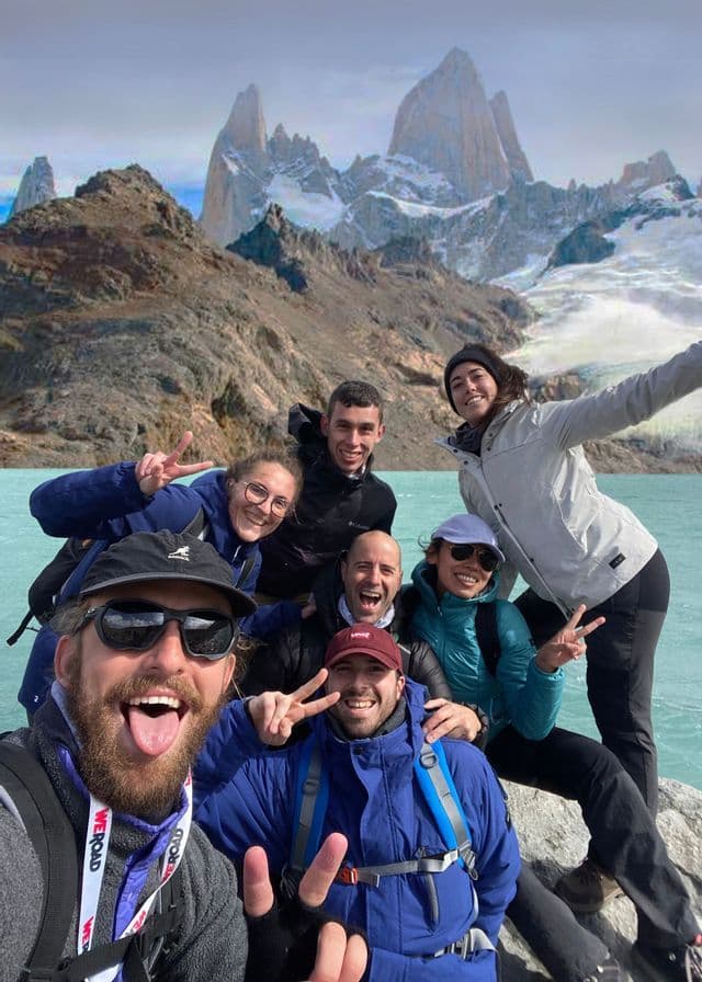 A WeRoad group trip takes a smiling selfie by a turquoise lake with a jagged, snowy mountain range in the background.
