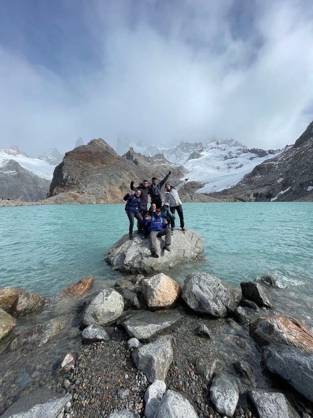 A WeRoad group trip posing together on a large rock in a turquoise alpine lake with snowy mountains behind them.