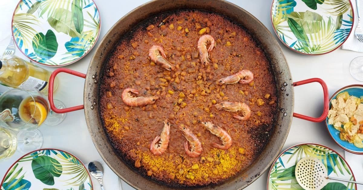 Draufsicht auf eine große Paellapfanne, gefüllt mit Reis und Garnelen, umgeben von Tellern mit Blattmuster und Gläsern.