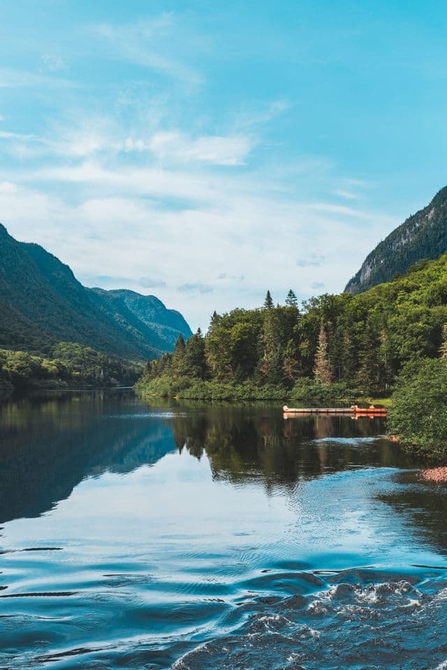 Un ampio fiume serpeggia attraverso una valle circondata da montagne boscose sotto un cielo azzurro, con due canoe a un molo.