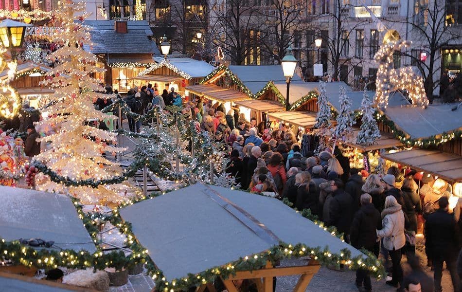 Luftaufnahme eines belebten Weihnachtsmarktes in der Dämmerung, mit Menschenmengen, die beleuchtete Holzstände und geschmückte Bäume bestaunen.