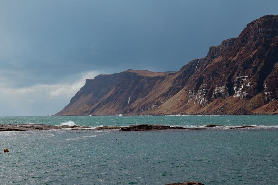 Une falaise brune et accidentée avec plusieurs cascades surplombe une mer turquoise agitée sous un ciel couvert.