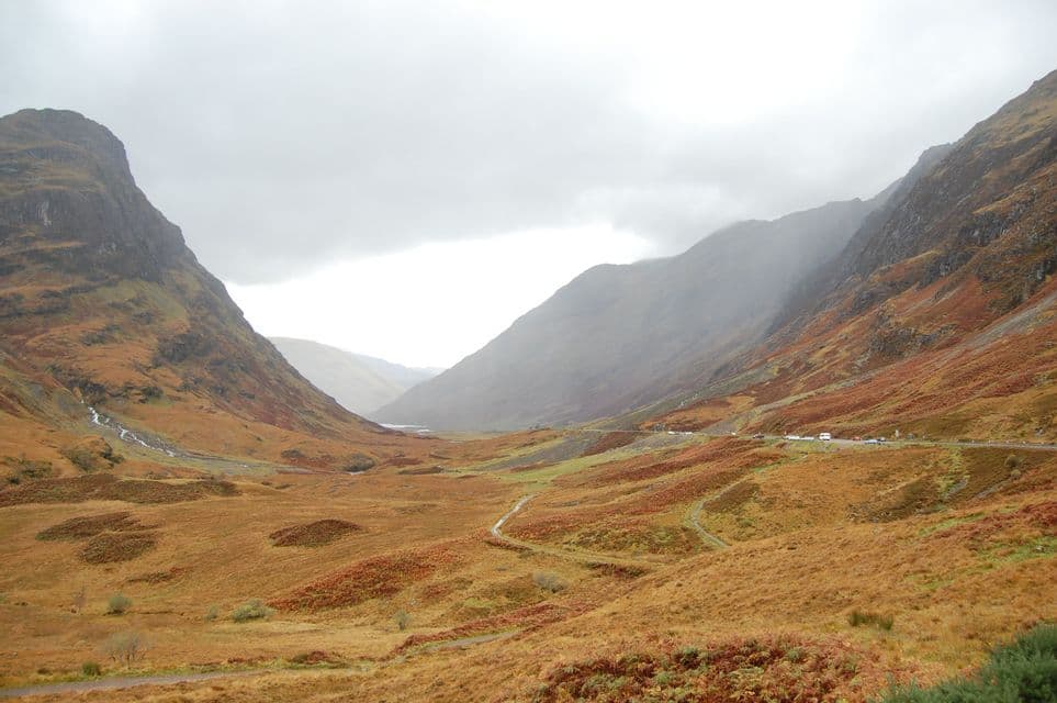 Une large vallée montagneuse avec des pentes couvertes de feuillage d'automne marron et orange sous un ciel couvert.