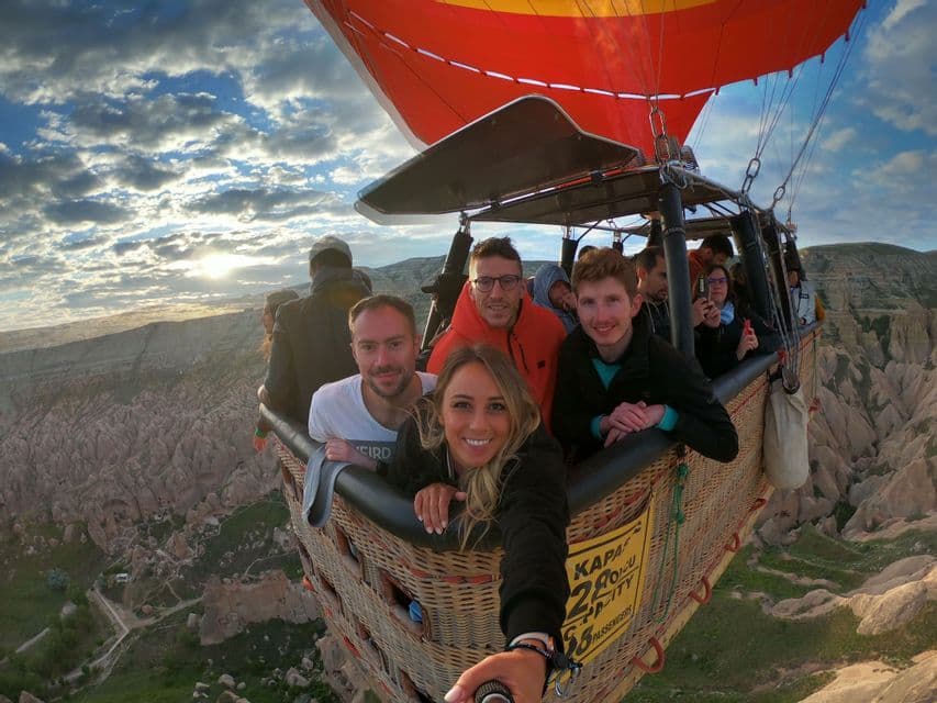Un grupo de WeRoad se toma una selfie desde la cesta de un globo aerostático volando alto sobre un paisaje rocoso al amanecer.