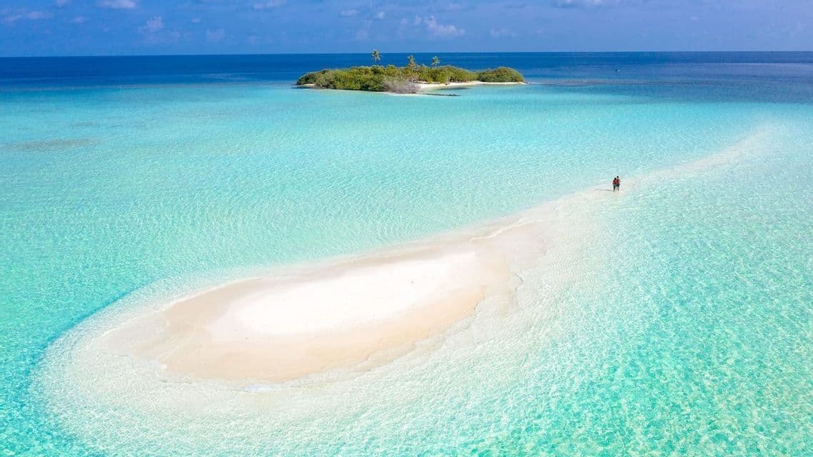 An aerial view of two people walking on a white sandbar that stretches through turquoise water towards a small, tropical island.