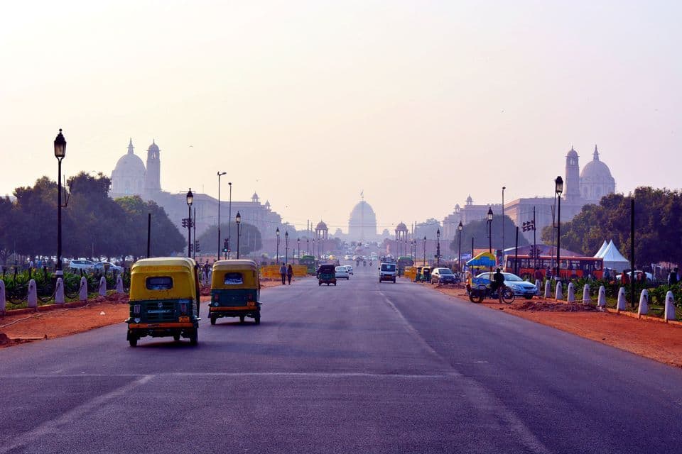 Due tuk-tuk guidano su un'ampia strada cittadina verso edifici monumentali con cupole sotto un cielo velato.