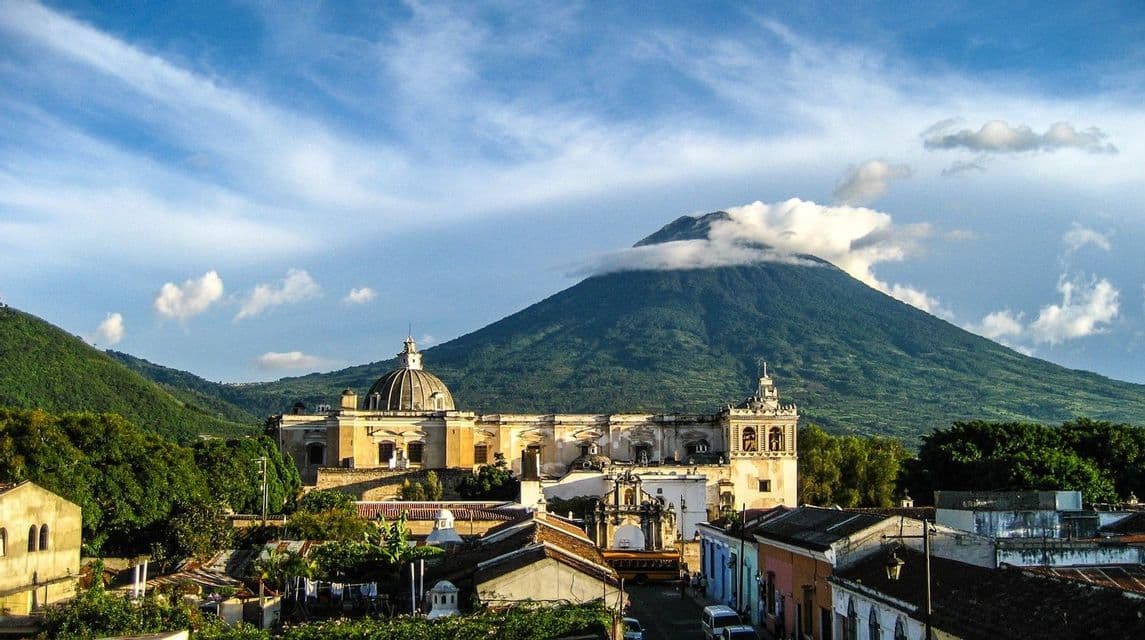 Vista sui tetti colorati della città verso una grande chiesa coloniale, con un enorme vulcano verde che si erge tra le nuvole alle sue spalle.