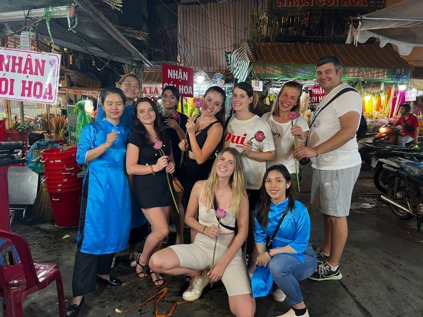 Eine WeRoad-Gruppenreise posiert für ein Foto auf einem Nachtmarkt, wobei viele Mitglieder rosa Lotusblumen halten.