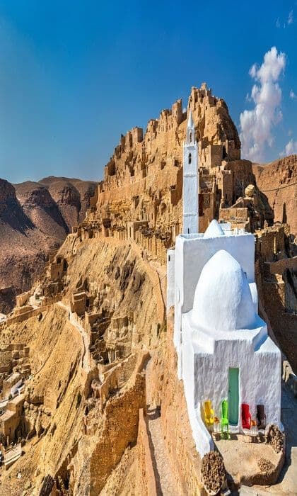 Una moschea imbiancata a calce con un minareto si erge su una scogliera, affacciata su un villaggio incastonato in una montagna rocciosa sotto un cielo azzurro.