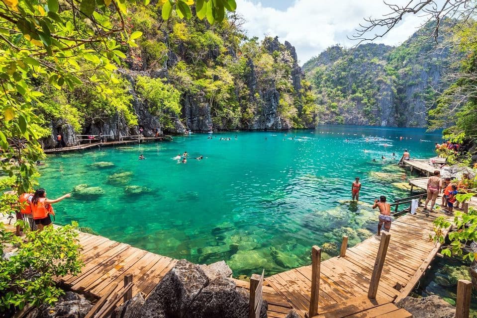 A WeRoad group trip swimming in a clear turquoise lagoon surrounded by rocky cliffs with lush greenery and wooden walkways.
