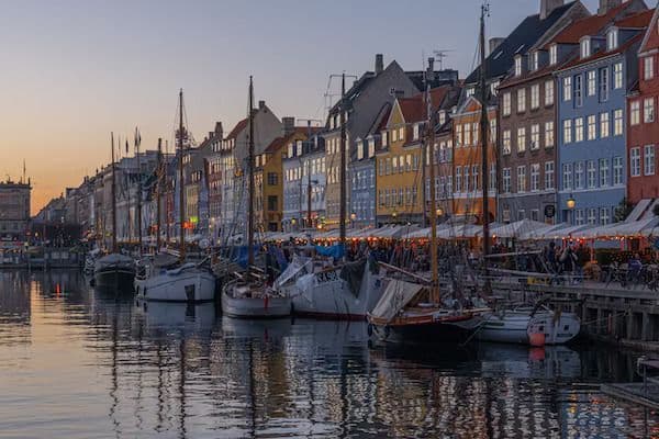 Barche a vela ormeggiate lungo un canale di fronte a edifici storici e colorati con caffè sul lungomare al tramonto.