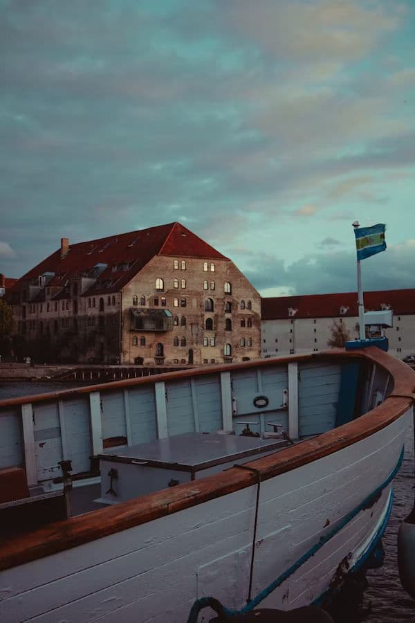 Ein Blick von einem Holzboot zeigt große, alte Backsteingebäude an einer Uferpromenade unter bewölktem Himmel in der Dämmerung.