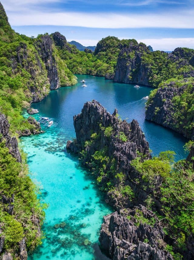 An aerial view of a tropical lagoon where boats float on turquoise water between steep, jungle-covered rocky cliffs.