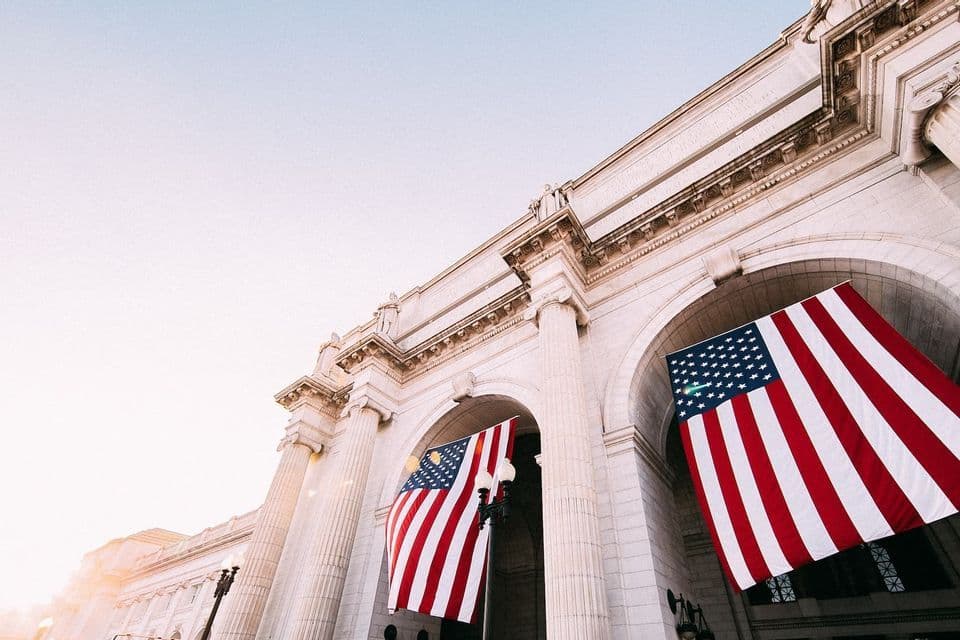 Una vista dal basso di due grandi bandiere americane appese agli archi di un classico edificio in pietra.