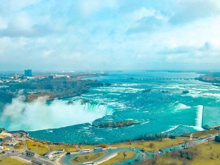 An aerial view of a large, horseshoe-shaped waterfall with vibrant turquoise water, a viewing area, and a distant cityscape.