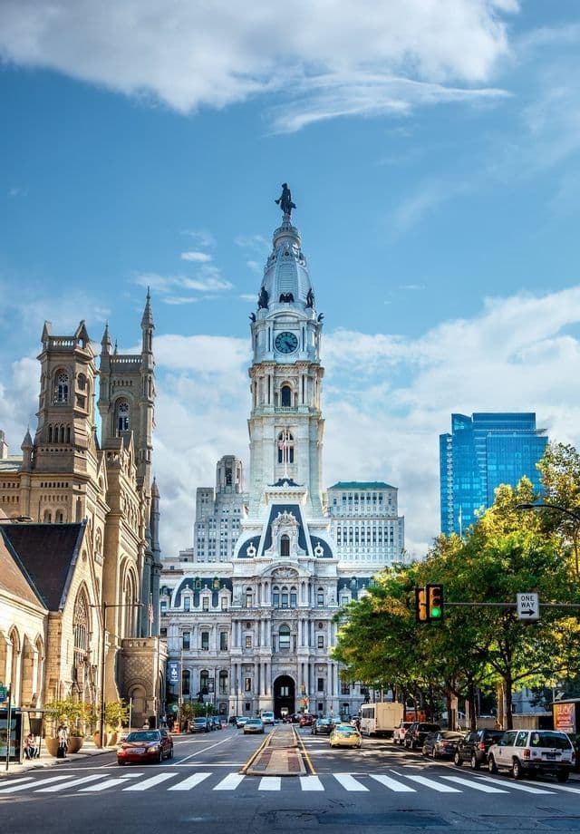 A grand historic building with a prominent clock tower stands at the end of a city street filled with cars and trees.