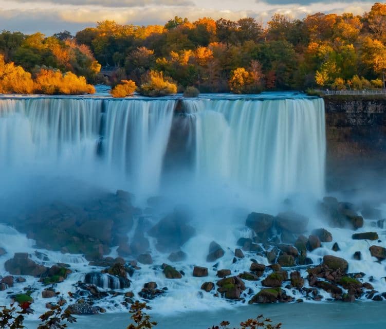A wide waterfall tumbles over a rocky ledge into a misty basin, backed by a forest with golden autumn leaves.