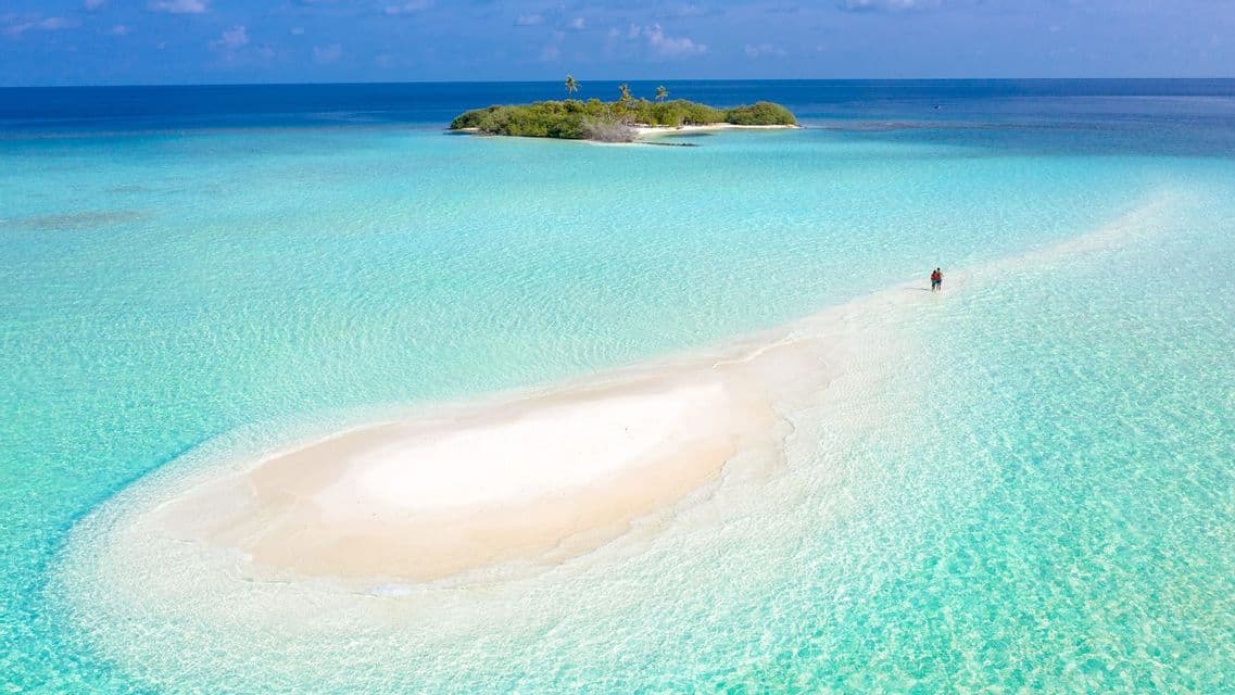 Vista aerea di due persone che camminano su una lingua di sabbia bianca in acqua turchese, con una piccola isola tropicale in lontananza.