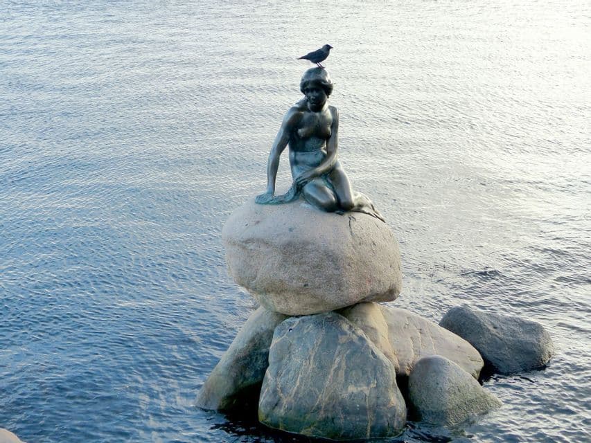 Eine Bronzestatue einer Meerjungfrau sitzt auf einem Felsen im Wasser, mit einem kleinen schwarzen Vogel auf ihrem Kopf.