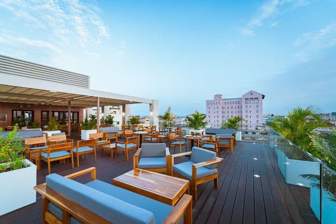 Una terrazza panoramica con mobili da esterno in legno e piante in vaso che si affaccia su uno skyline cittadino sotto un cielo blu.