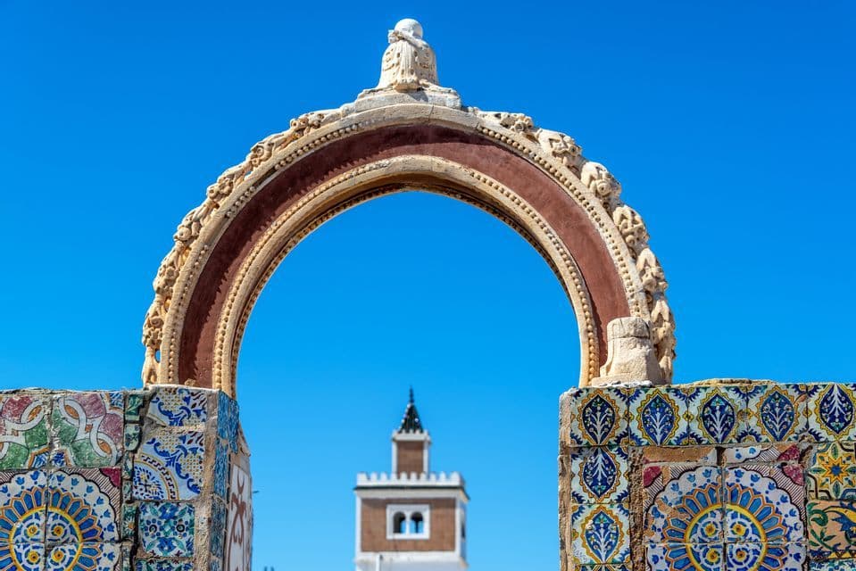 Un arco ornato con piastrelle colorate incornicia una torre distante sotto un cielo azzurro chiaro.
