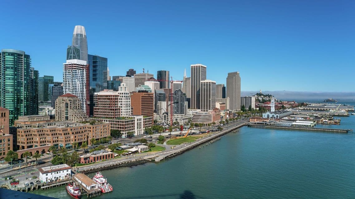 Vista panoramica diurna di uno skyline moderno con grattacieli e un molo sul lungomare sotto un cielo azzurro e limpido.