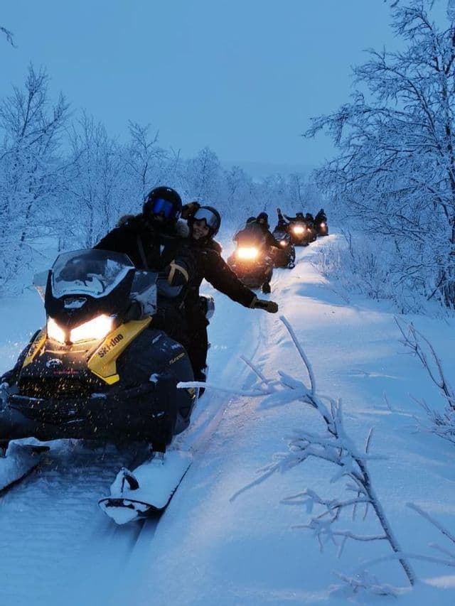 A WeRoad group trip riding snowmobiles with headlights on along a snowy path through a forest at dusk.