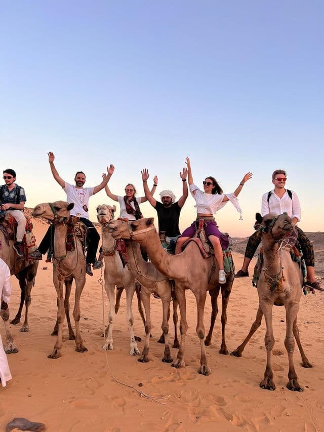 A WeRoad group trip of people smiling and waving their hands while riding camels in a desert at dusk.