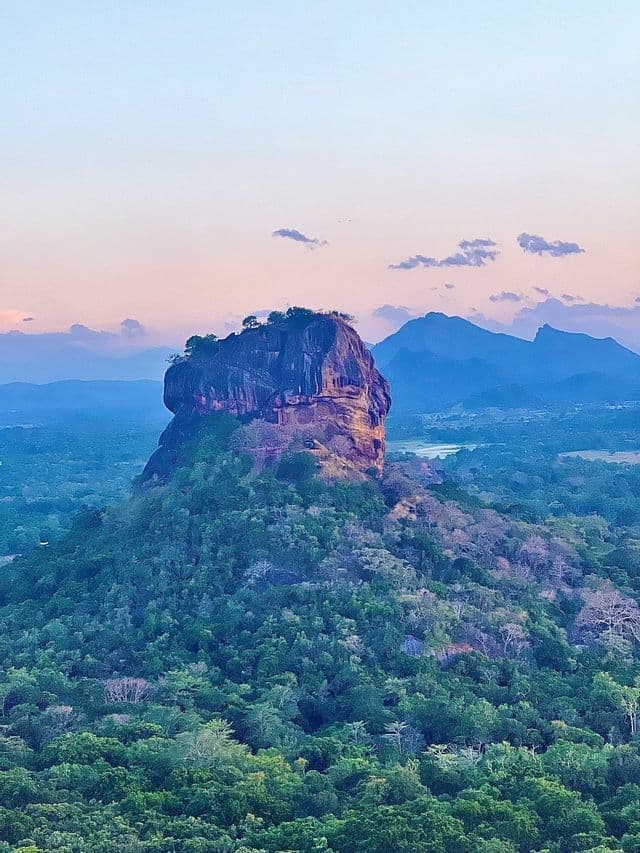 Una imponente formación rocosa con árboles en la cima se eleva sobre una densa selva verde, con montañas distantes bajo un cielo de tonos pastel al atardecer.
