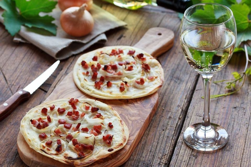 Two Alsatian Flammkuchen with bacon and onions on a wooden board next to a glass of white wine on a rustic table.