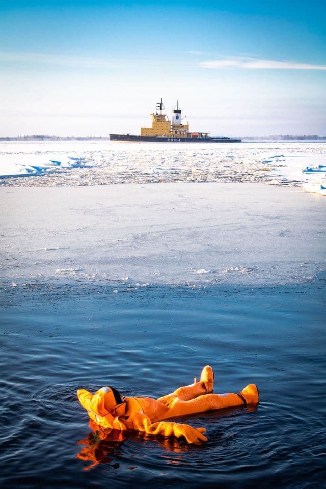 Une personne en combinaison de survie orange vif flotte dans l'eau glacée avec un brise-glace en arrière-plan.