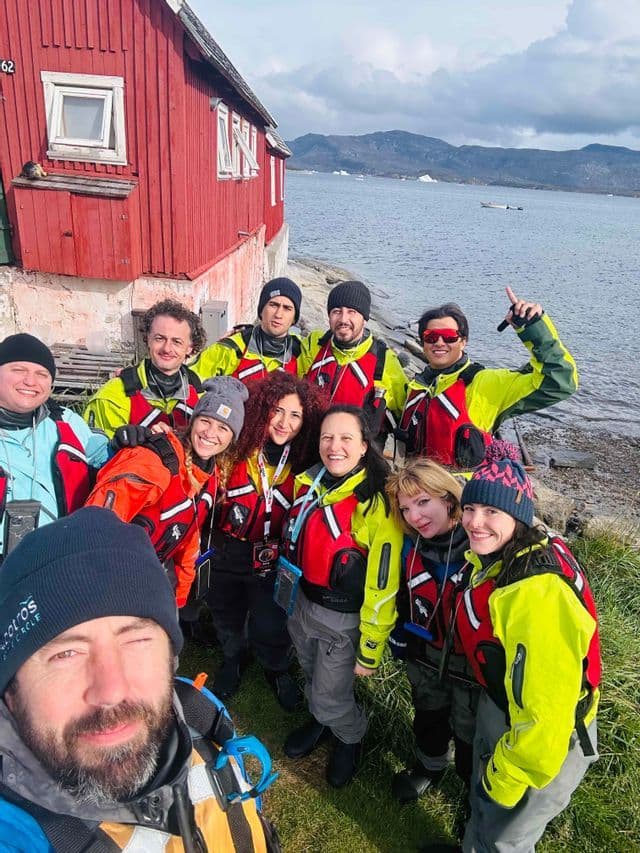 Un gruppo WeRoad in gilet di salvataggio posa per un selfie su una riva erbosa accanto a un edificio rosso.