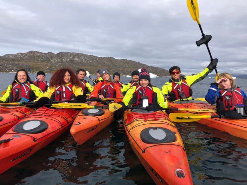 Un gruppo WeRoad in kayak arancioni sull'acqua si fa fotografare, con una costa rocciosa e iceberg sullo sfondo.