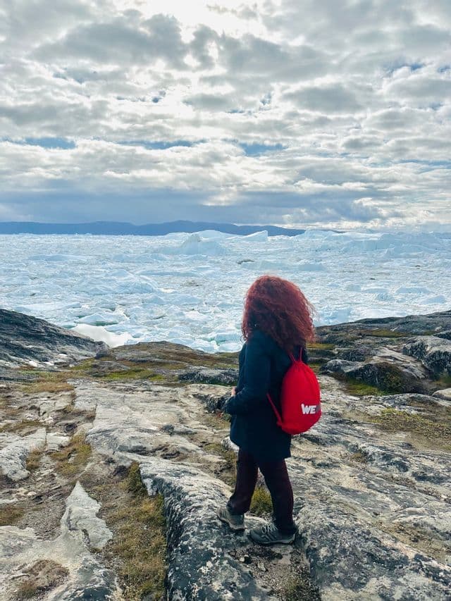 Una persona dai capelli rossi con uno zaino WeRoad rosso è in piedi su una costa rocciosa, ammirando un mare di iceberg sotto un cielo nuvoloso.