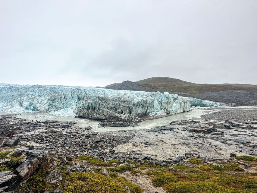 Un'ampia parete glaciale con un fiume di acqua di disgelo che scorre dalla sua base attraverso un paesaggio roccioso con rada vegetazione sotto un cielo nuvoloso.