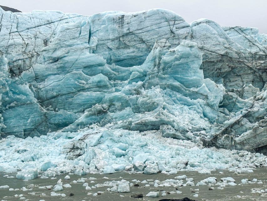 La parte terminale di un grande ghiacciaio, con ghiaccio blu frastagliato e scure linee di sedimento, che si frantuma nell'acqua.