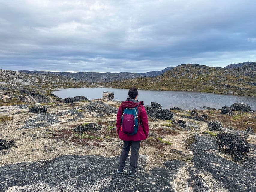 Una persona con una giacca rossa e uno zaino sta su un terreno roccioso, guardando un lago circondato da colline sotto un cielo nuvoloso.