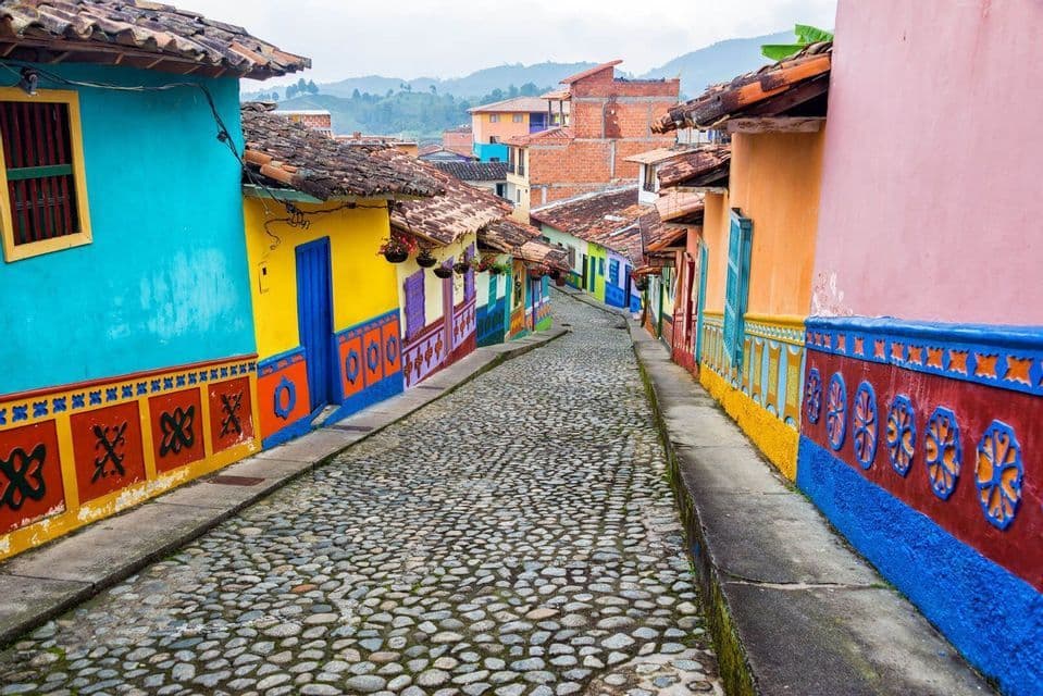 An empty cobblestone street lined with brightly colored houses with tiled roofs, winding uphill towards a distant mountain range.