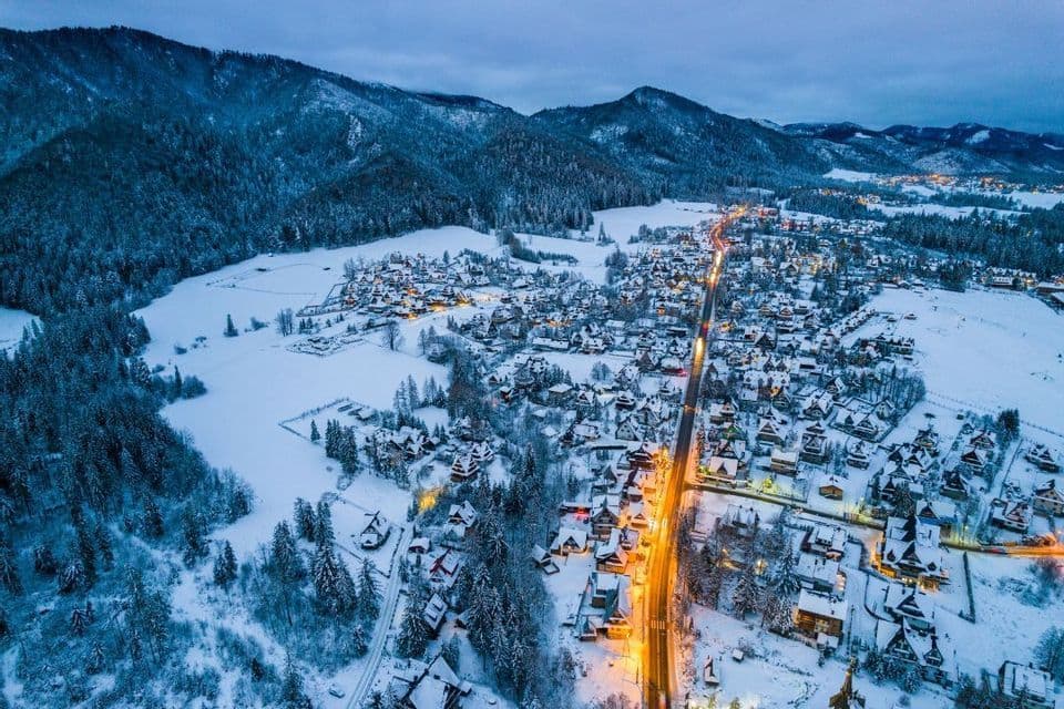 Vista aerea di un villaggio immerso in una valle montana innevata al crepuscolo, con strade e case illuminate.