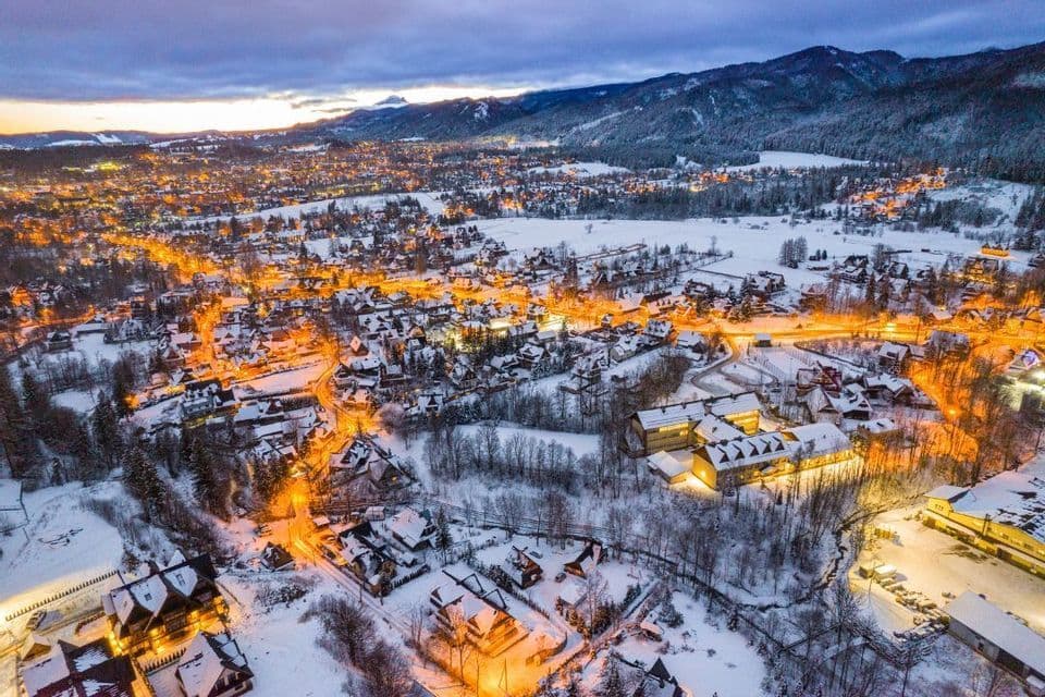 Una vista aerea di una cittadina innevata illuminata dalle luci al crepuscolo, con una catena montuosa sullo sfondo.