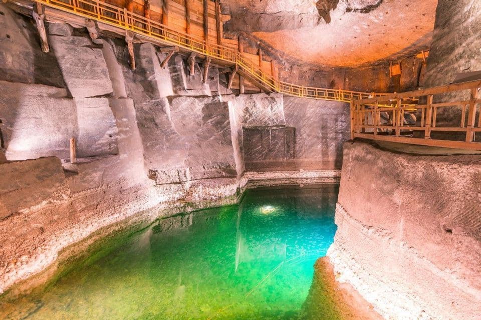 Una grande caverna illuminata con passerelle di legno incassate nelle pareti rocciose sopra una pozza d'acqua verde.