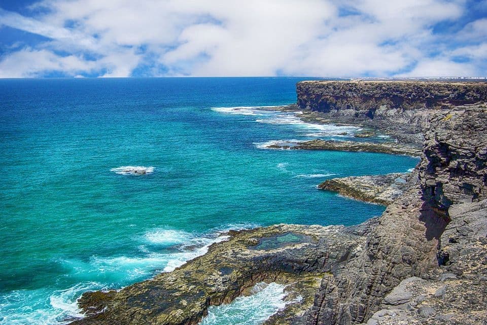 Una costa frastagliata e rocciosa costeggia un oceano turchese con piccole onde bianche sotto un cielo azzurro con nuvole soffici.