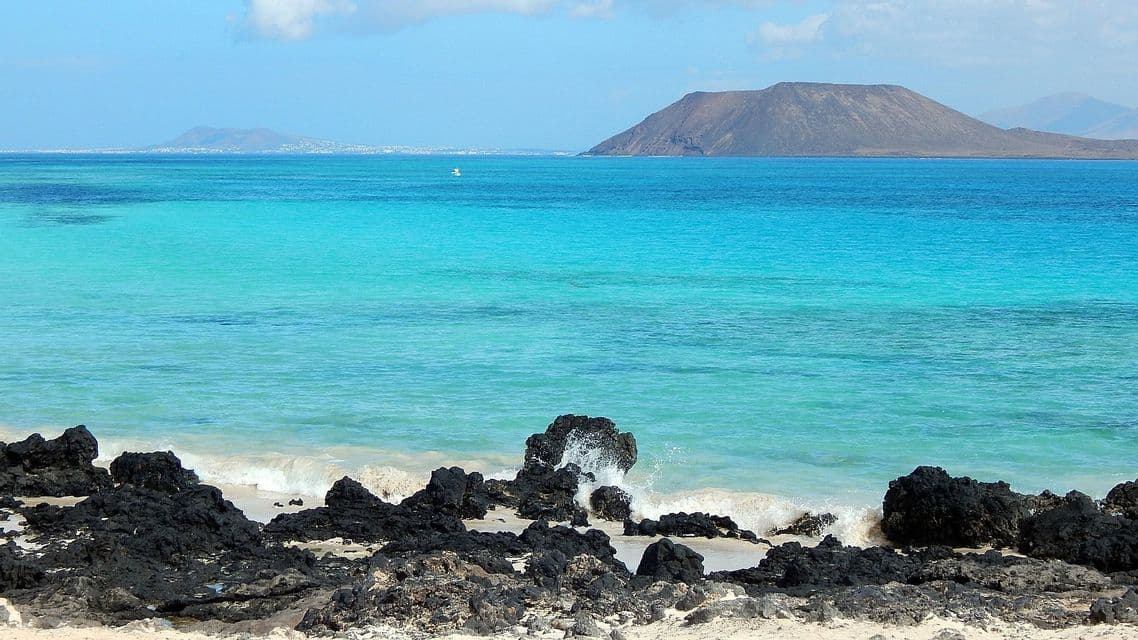 Acqua turchese lambisce rocce vulcaniche nere su una spiaggia sabbiosa, con un'isola vulcanica in lontananza sotto un cielo blu.