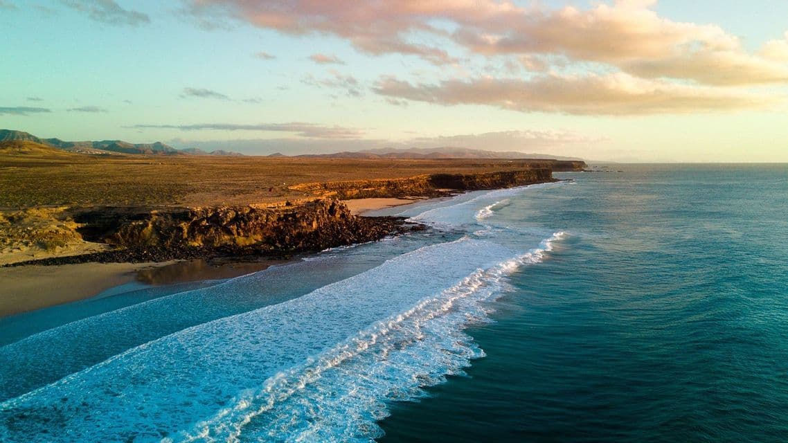 Vista aerea di una costa frastagliata dove le onde turchesi dell'oceano incontrano una spiaggia sabbiosa e scogliere aride al tramonto.