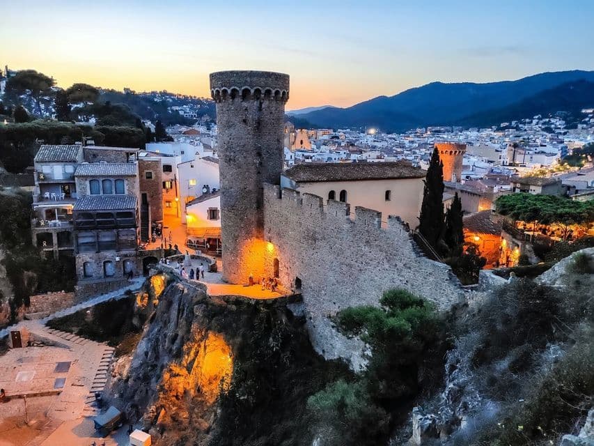 Un castello di pietra con una torre si erge illuminato su una scogliera che domina una città e le montagne al tramonto.